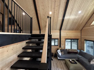 Open black timber staircase beside a finished living room with wood walls, vaulted ceiling, and dark structural beams
