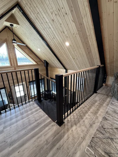 Finished timber loft stair landing with black railings, natural wood handrails, vaulted wood ceiling, and triangular windows