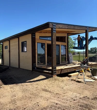 Modern low-slope timber home under construction with natural wood siding, dark structural posts, tall windows, and covered deck