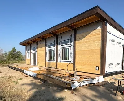 Modern timber home exterior during siding installation with dark structural framing, large windows, and a low-slope roof