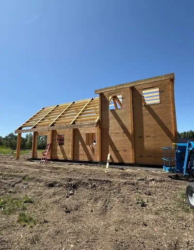 Timber home under construction with wall sheathing, roof framing, window openings, and lift equipment under a clear blue sky