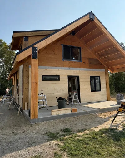 Timber home nearing completion with natural wood siding, dark roof and trim, covered entry, and large black-framed windows