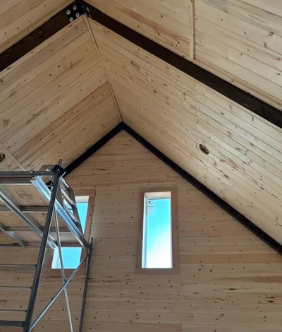 Interior finishing work on a vaulted timber ceiling with dark structural beams, narrow windows, and scaffold