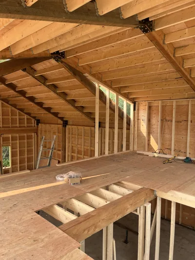 Interior construction view from an unfinished loft showing timber roof framing, floor framing, structural beams, and wall studs