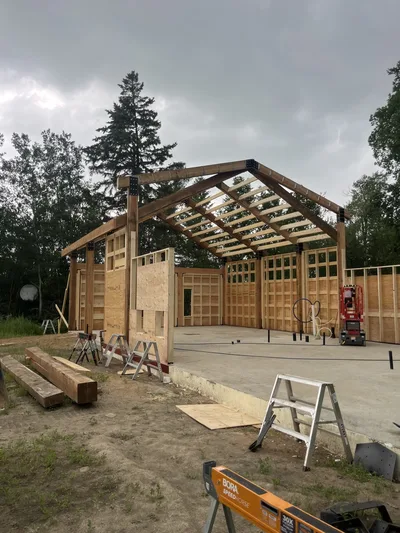 Timber home under construction with large structural posts and beams rising above framed walls on a concrete slab