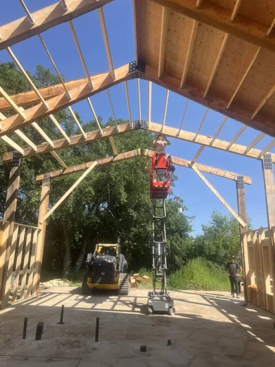 Interior construction view looking up through exposed timber beams and roof framing while a lift is in use