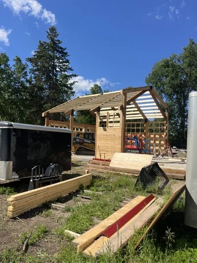 Side view of a timber home under construction with exposed roof framing, wall framing, equipment, and materials