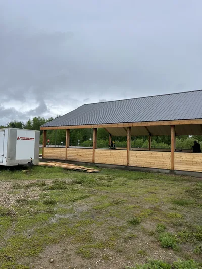 Open-sided timber pavilion with natural wood infill walls, exposed posts, and a dark metal roof