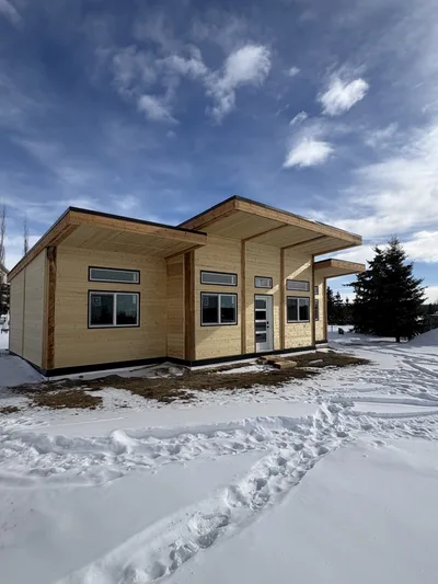 Modern timber home in winter with natural wood siding, black trim, large windows, and broad roof overhangs