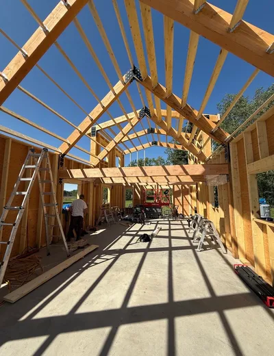 Interior construction view of exposed timber roof framing, beams, wall framing, and strong roof-shadow patterns