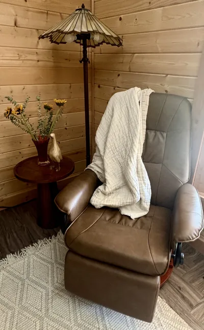 Leather recliner beside a wood side table, flowers, and floor lamp against timber-finished walls
