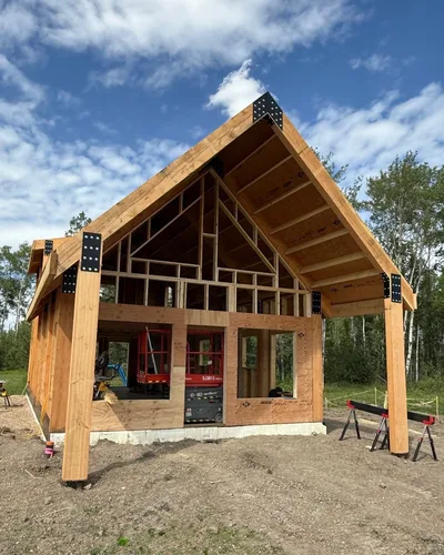 Front view of a timber home under construction with dramatic gable framing, exposed connectors, and framed wall openings