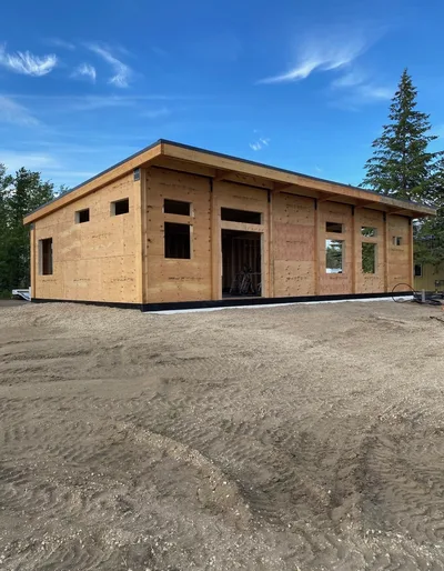 Low-slope timber building shell under construction with sheathed walls, window openings, and deep roof overhang