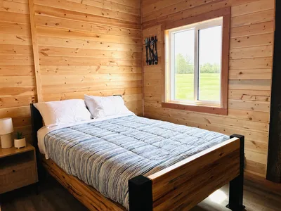 Finished timber bedroom with natural wood walls, wood-and-black bed frame, and a large window overlooking a green field