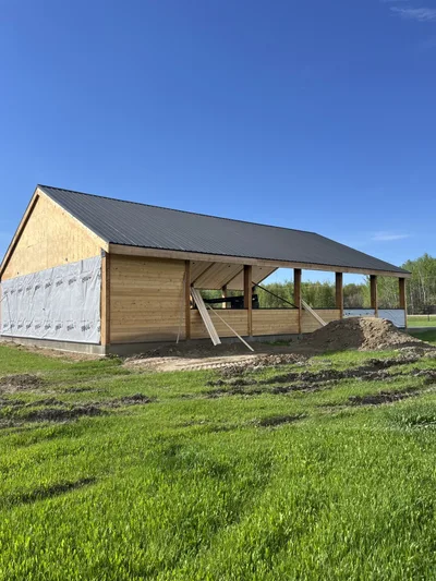 Open timber pavilion under construction with dark metal roof, partial wood walls, and natural timber posts