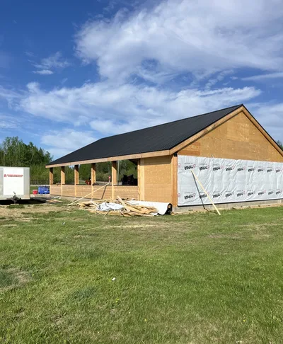 Timber pavilion under construction with dark metal roof, partial wall sheathing, open bays, and a Timber Built Homes trailer nearby