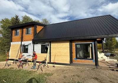 Timber home exterior during finishing with black roof and trim, natural wood siding, and workers installing cladding